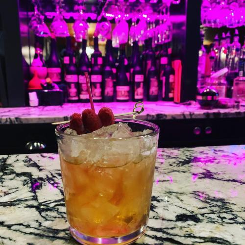 A cocktail with raspberries on top sits on a marble bar, illuminated by pink lights and a blurred bar backdrop.