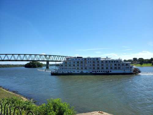 A large riverboat sails under a bridge on a sunny day, surrounded by calm waters and greenery.