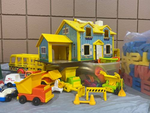 A colorful toy house surrounded by various toy vehicles and construction equipment on a gray surface.