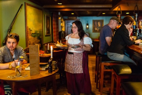 A waitress in a traditional outfit serves food in a cozy restaurant with patrons at the bar and tables.