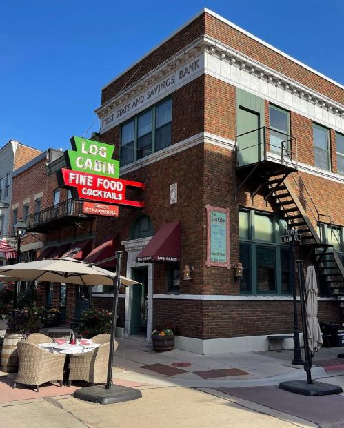 A brick building with a sign for "Log Cabin Fine Food" and outdoor seating, located on a sunny street.
