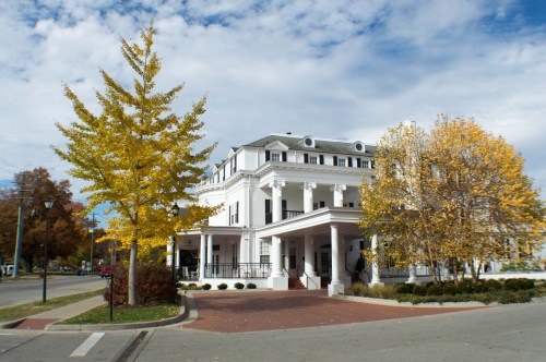 A large white building with columns, surrounded by autumn trees with yellow leaves under a partly cloudy sky.