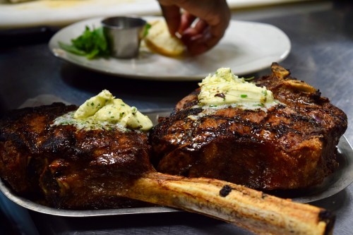 Two grilled ribeye steaks topped with herb butter, served on a metal platter with a side of garnishes.