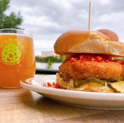A crispy chicken sandwich with pickles and crushed chips, alongside a glass of beer, set against a cloudy backdrop.