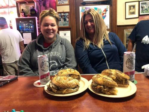 Two women sit at a bar with bagels and drinks, smiling at the camera. Plates with food are in front of them.