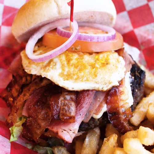 A hearty burger topped with bacon, ham, a fried egg, lettuce, tomato, and onion, served with crinkle-cut fries.