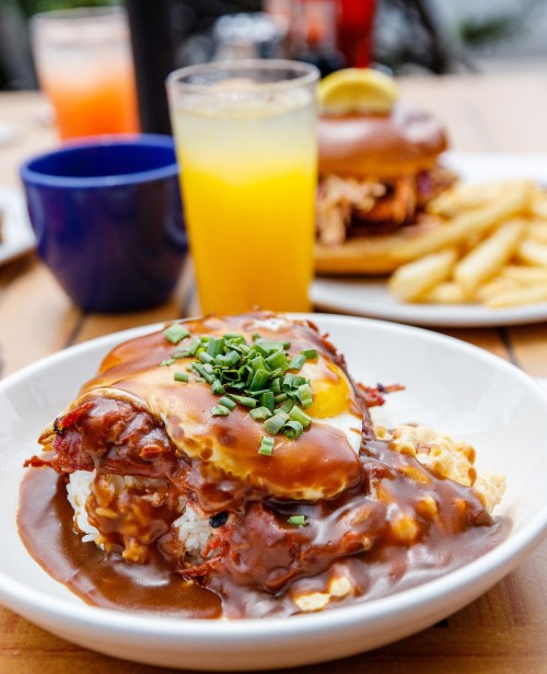 A plate of rice topped with a fried egg, savory sauce, and green onions, with drinks and a burger in the background.