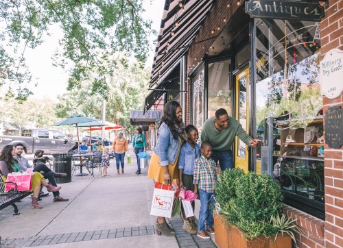 A family admires items in a shop window while shopping in a vibrant outdoor area with other people nearby.