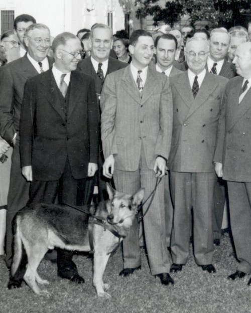 A group of men in suits stands together outdoors, with a dog on a leash in front of them.