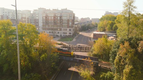 A train crosses a bridge over a road, surrounded by trees and buildings in a sunny urban setting.