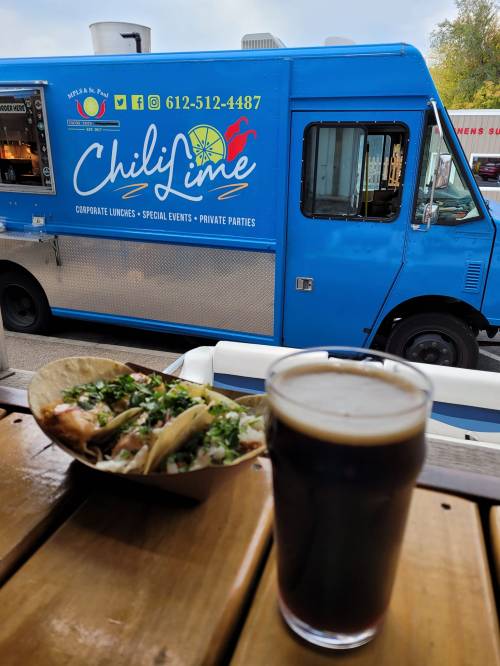 A blue food truck named "Chili Lime" parked beside a wooden table with tacos and a glass of dark beer.
