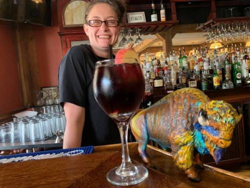 A smiling bartender stands behind a bar with a colorful bison figurine and a glass of sangria.