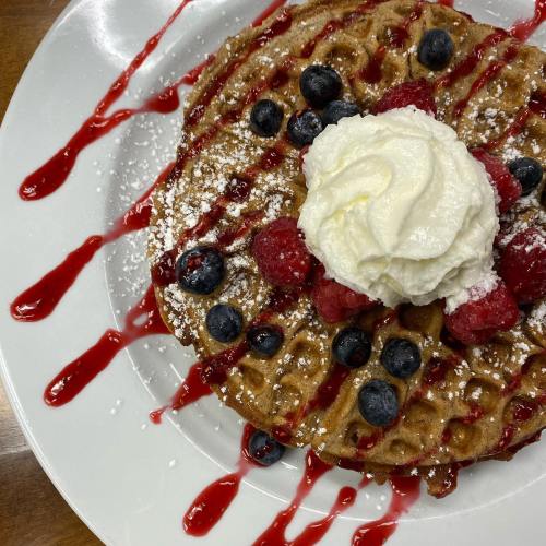 A delicious waffle topped with whipped cream, raspberries, blueberries, and drizzled with raspberry sauce.