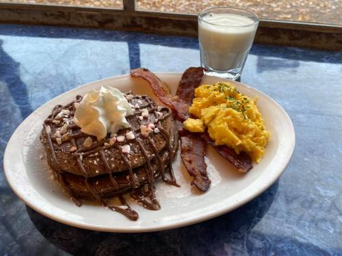 A plate with chocolate pancakes topped with whipped cream and peppermint, scrambled eggs, bacon, and a glass of milk.