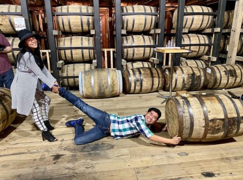 A woman playfully pulls a man lying on the floor next to wooden barrels in a rustic setting.