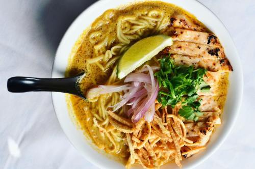 A bowl of ramen with grilled chicken, lime, onions, cilantro, and crispy noodles in a rich broth.