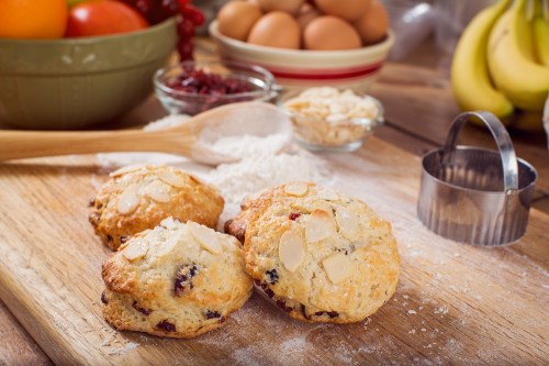 Freshly baked scones with almonds on a wooden surface, surrounded by baking ingredients like flour, eggs, and fruit.