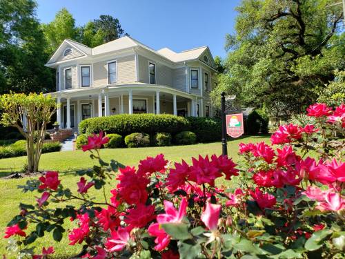 A charming two-story house surrounded by lush greenery and vibrant pink roses in the foreground.