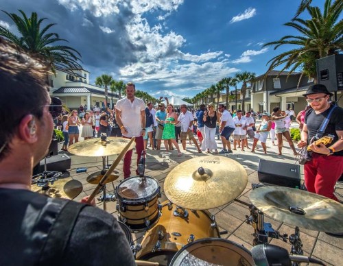 A lively outdoor concert scene with a drummer in the foreground and a crowd dancing under palm trees.
