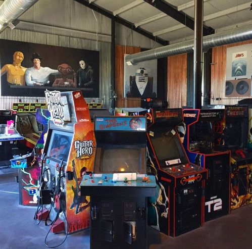 A vibrant arcade filled with classic arcade machines, including Guitar Hero and various retro games, in a spacious venue.
