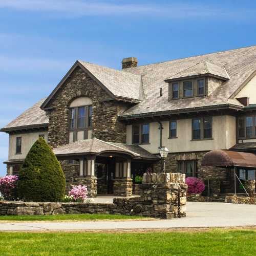 A large, elegant stone house with a steep roof, surrounded by greenery and colorful flowers on a sunny day.