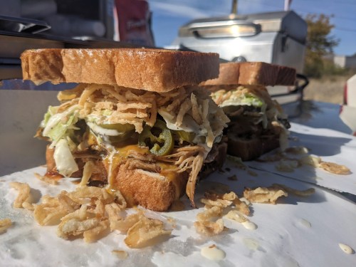 A close-up of a sandwich with pulled pork, lettuce, jalapeños, and crispy onions between two slices of bread.