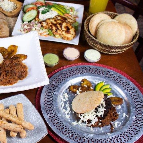 A table spread with various dishes, including grilled chicken, salads, plantains, and tortillas on a decorative plate.