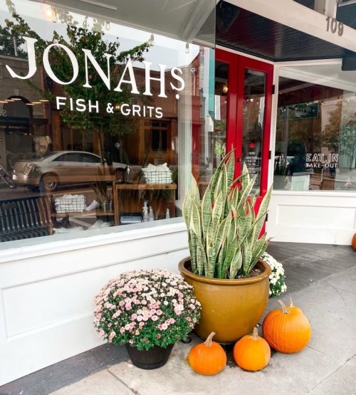 A storefront with "JONAH'S FISH & GRITS" sign, featuring potted plants, flowers, and pumpkins on the sidewalk.