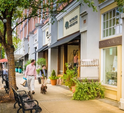 A couple walks a dog along a tree-lined street with shops and outdoor seating.