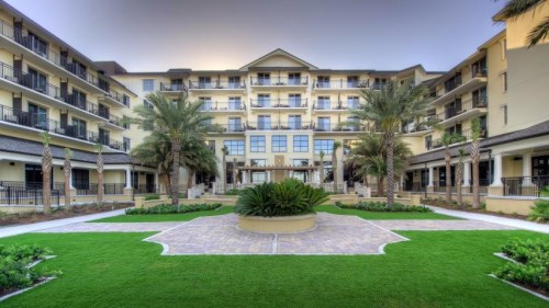 A modern hotel with multiple floors, surrounded by palm trees and landscaped gardens in a sunny setting.