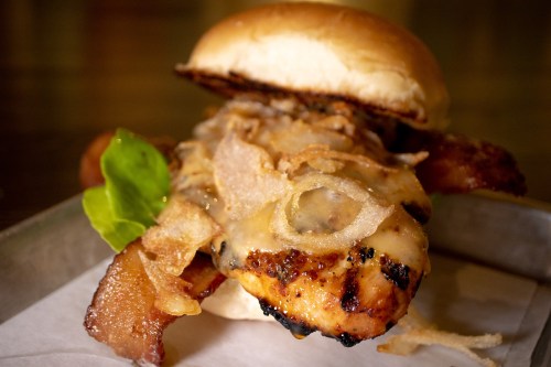 A close-up of a grilled chicken sandwich topped with bacon, melted cheese, crispy onions, and lettuce on a soft bun.