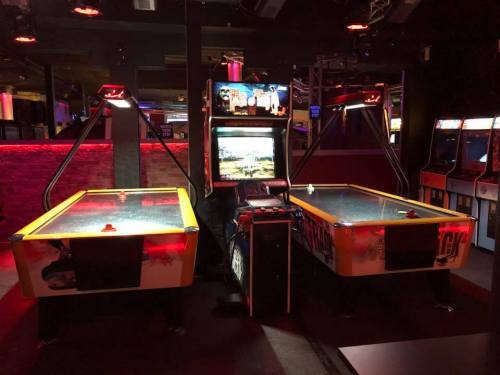 Two air hockey tables in a dimly lit arcade, with colorful lights and a vintage arcade game in the background.
