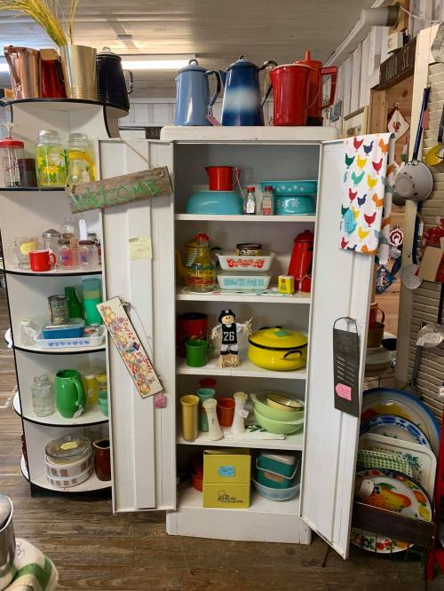 A vintage cupboard filled with colorful kitchenware, including pots, bowls, and decorative items, in a cozy shop setting.