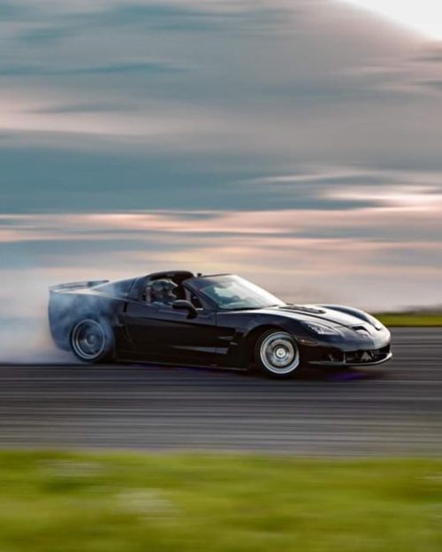 A black sports car drifts on a racetrack, leaving a trail of smoke against a cloudy sky.