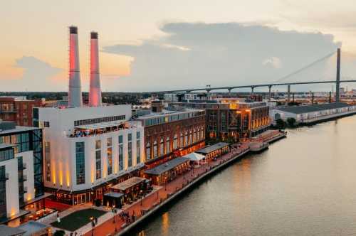 A waterfront view of a cityscape at sunset, featuring a building with two smokestacks and a bridge in the background.