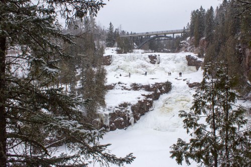 A snowy landscape featuring a frozen waterfall and a bridge in the background, surrounded by evergreen trees.