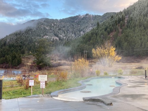 A serene hot spring pool surrounded by mountains and autumn foliage, with steam rising in the cool air.