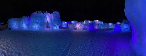 A panoramic view of an ice sculpture park illuminated with colorful lights at night, surrounded by snow.