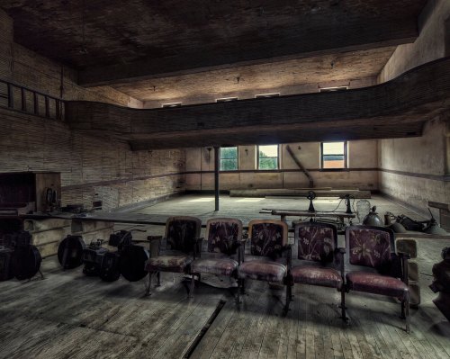 Abandoned interior of a spacious room with worn chairs, wooden floors, and large windows letting in natural light.