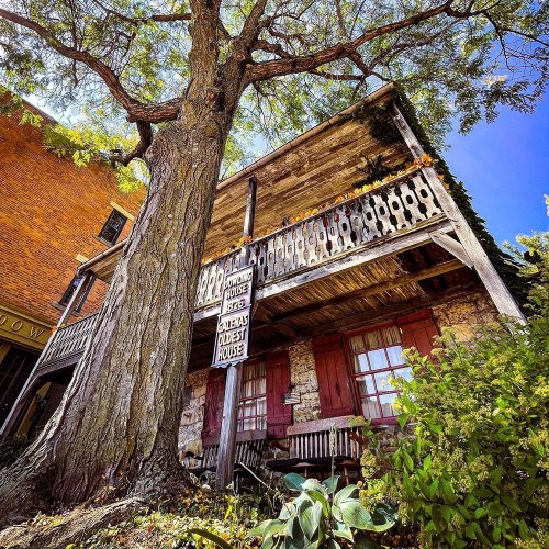 A historic wooden house with a balcony, surrounded by a large tree and greenery, labeled as "America's Oldest House."