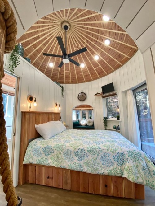 Cozy circular bedroom with a wooden ceiling, large bed, and natural light from windows, featuring plants and modern decor.