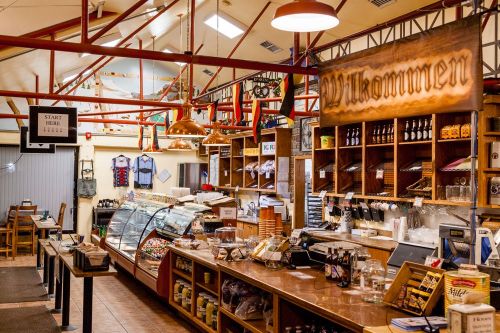 A cozy café interior with wooden shelves, a display case, and a "Willkommen" sign, featuring warm lighting and rustic decor.