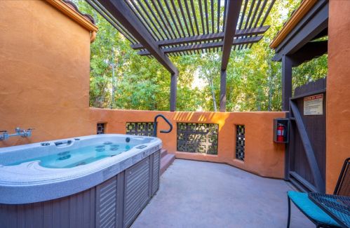 A private outdoor hot tub surrounded by greenery and a wooden pergola, with a safety sign and a chair nearby.