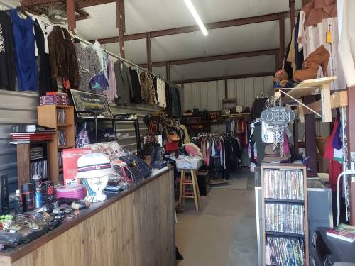 Interior of a thrift store featuring clothing racks, shelves of items, and a checkout counter. Sign reads "OPEN."