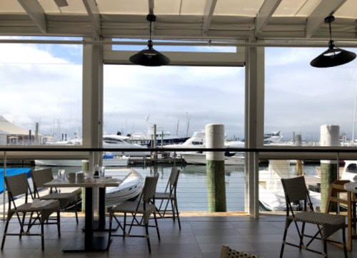 View from a waterfront restaurant showing empty tables and boats docked in the marina under a cloudy sky.