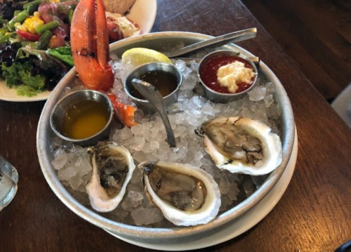 A seafood platter featuring oysters on ice, a lobster tail, lemon, and dipping sauces.