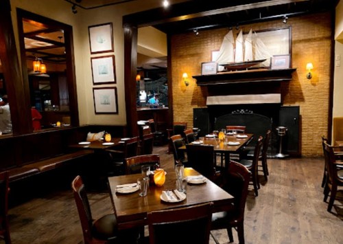 Cozy restaurant interior with wooden tables, chairs, and a fireplace, featuring nautical decor and warm lighting.