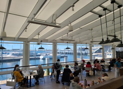 A bright restaurant interior with diners, large windows overlooking a marina filled with boats.