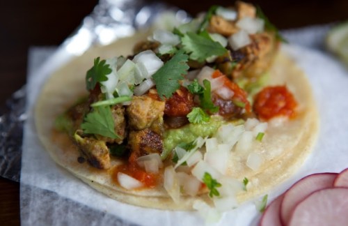 A close-up of a taco filled with grilled chicken, guacamole, salsa, onions, and cilantro on a soft tortilla.