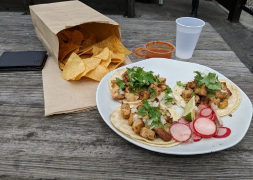 A plate of tacos with grilled chicken, cilantro, and radishes, alongside a bag of tortilla chips and a cup of salsa.
